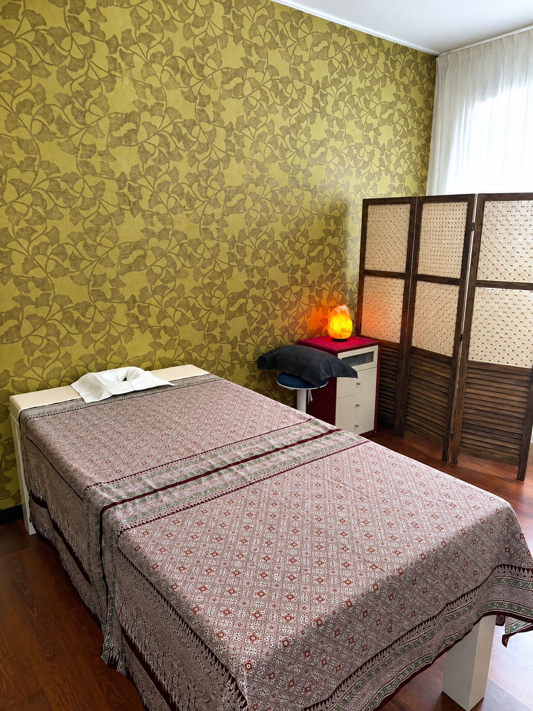 De behandelkamer met gouden ginkgo-behang, een opgemaakt massagebed en een zoutlamp.