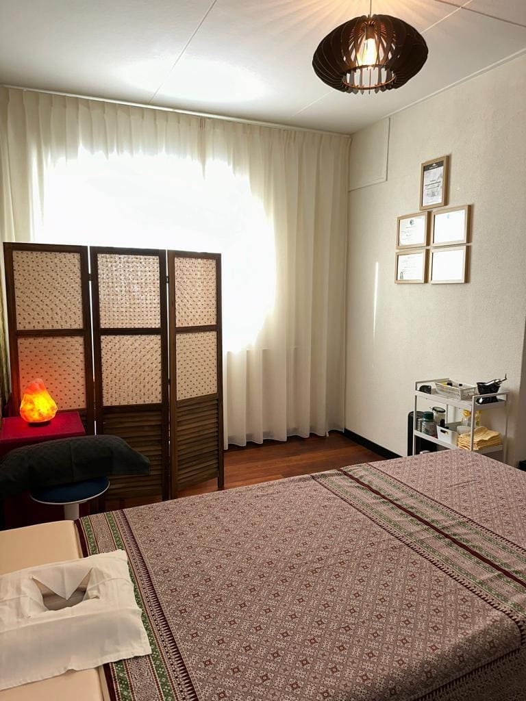 Daglicht door de vitrage in de behandelkamer, met een opgemaakt bed en een zoutlamp.